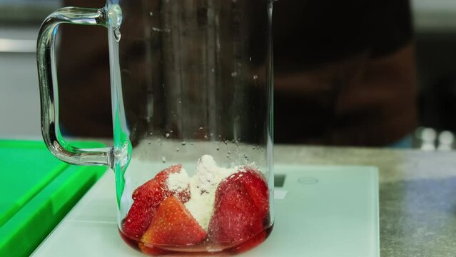 Sprinkling sugar powder into transparent jug with strawberries standing on scales. Measuring required amount of ingredients closeup