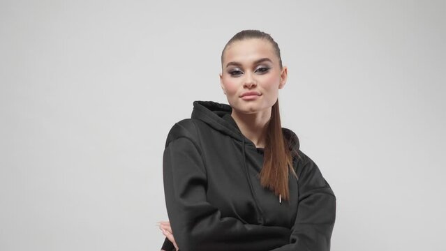 A young female model with bold makeup, winged eyeliner and a stylish ponytail hairstyle in a fashionable youth black hoodie smiles and poses in the studio on a white background, looking at the camera.