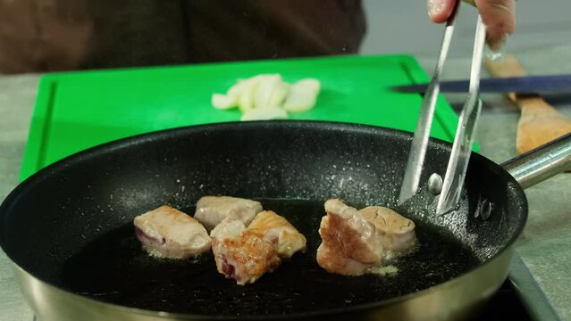 Hand of professional chef with tongs turns over frying slices of meat. Beef cubes get appetizing golden crust on all sides