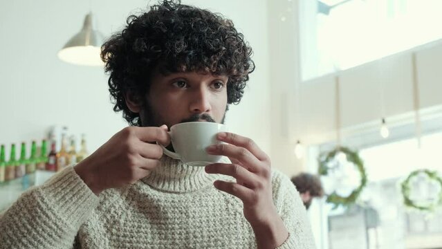 Relaxed curly haired indian man in beige sweater drinking coffee in cafe