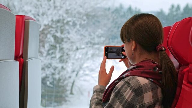 Young woman takes a photo of the winter landscape outside the window while sitting inside a public transport. The tourist train makes stops and a passenger takes photos with his phone