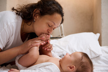 Loving caring mom gently kisses little feet, tiny toes of her newborn baby boy, enjoys spending...