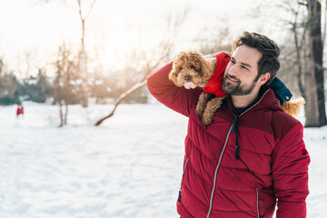 Handsome man holding dog poodle on his shoulder while walking outdoors in the snow enjoying in...