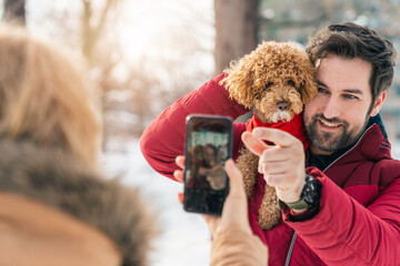 Adult couple taking picture with poodle dog outdoors. Woman photographing with mobile phone man and...