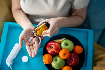 Focus on the hands of unrecognizable sick woman taking medications, lying in bed with tray table with fresh fruits, ear thermometer and tea mug. - obrazy, fototapety, plakaty