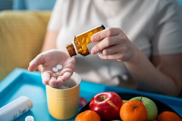 Focus on the hands of unrecognizable sick woman taking medications from pill bottle, sitting at home with tray bed table with fresh fruits, ear thermometer and tea mug on it. Taking medicine with tea.