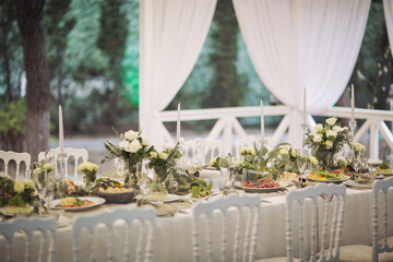 Stylish elegant table setting for festive dinner. The chairs and table for guests, served with cutlery, greenery flowers and crockery and covered with a white tablecloth. Catering on Wedding. Banquet.