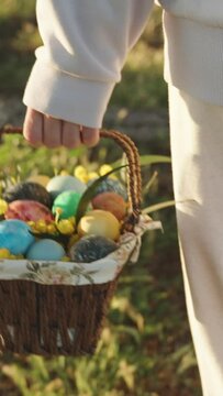 Vertical video. A walk behind a woman in white carrying a basket of painted Easter eggs. Sunny forest at dawn.