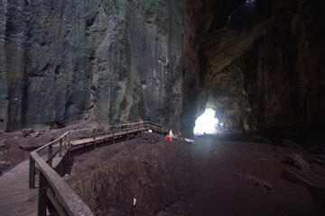A person exploring a spacious cave with a wooden walkway leading towards the illuminated cave...