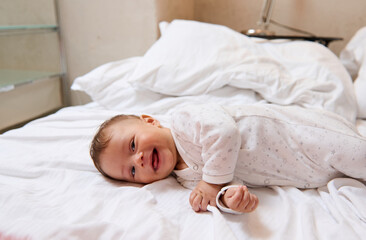 Fototapeta na wymiar Playful lovely baby boy in white clothes, smiling and laughing lying on white bedsheets on the bed at home