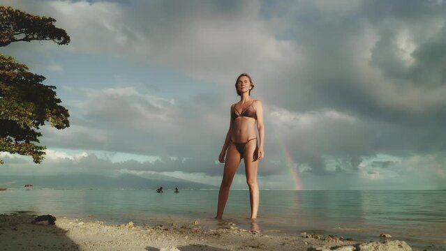 Woman in bikini walking tropical beach under a rainbow sky. Dramatic evening sunset weather after rain storm. Outdoor lifestyle travel on summer holiday vacation. Beautiful nature seascape landscape