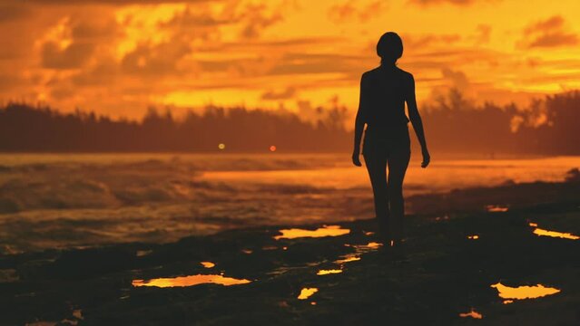 Woman silhouette walk coastline in orange sunset. Slim female enjoy evening bright sky, stone beach, ocean waves. Stunning sunrise wild nature. Outdoor travel on summer holiday vacation. Slow motion