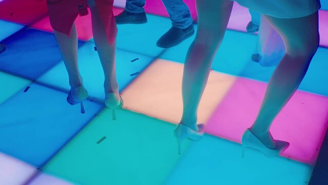 Girls in evening dresses and shoes with heels dance on luminous square disco floor. Vintage disco party with dancing girls. Party at nightclub in style of 80s. Colored flashing floor in nightclub