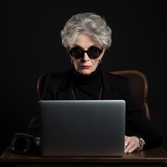Portrait of senior woman wearing sunglasses and black background sitting on a chair working with a laptop.