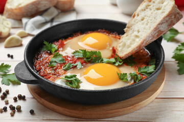 Delicious shakshuka with bread in frying pan on light wooden table, closeup