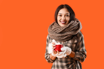 Beautiful young Asian woman in warm scarf holding cup of cocoa with marshmallows on orange...