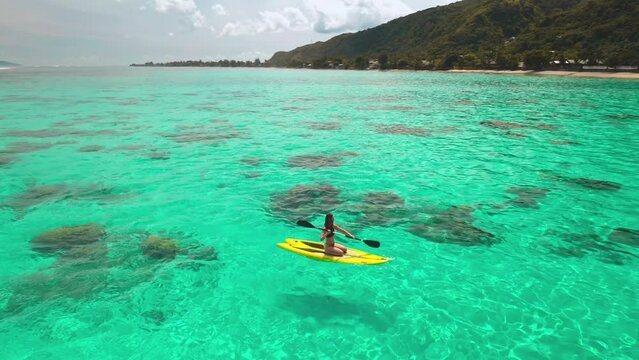 Aerial drone shot woman paddle boarding coral reef tropical island. Young female rows sitting on yellow paddleboard in crystal turquoise sea water. Outdoor lifestyle travel on summer holiday vacation.