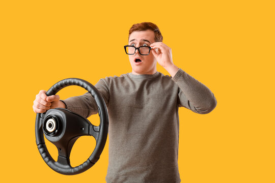 Shocked young man in eyeglasses with steering wheel on yellow background