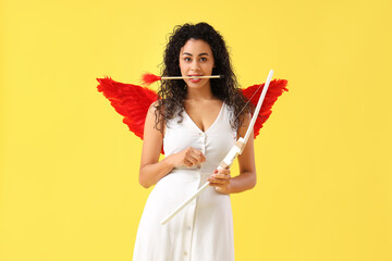 Young African-American woman dressed as Cupid with bow and arrow on yellow background. Valentine's...
