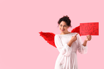 Young African-American woman dressed as Cupid holding paper with text I LOVE YOU on pink...