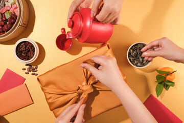 One person is wrapping a gift with a yellow silk cloth, one person is pouring tea, the other person...