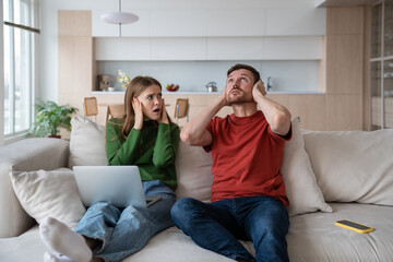 Exhausted irritated couple sitting on sofa with ears closed because of unpleasant nervous sound,...