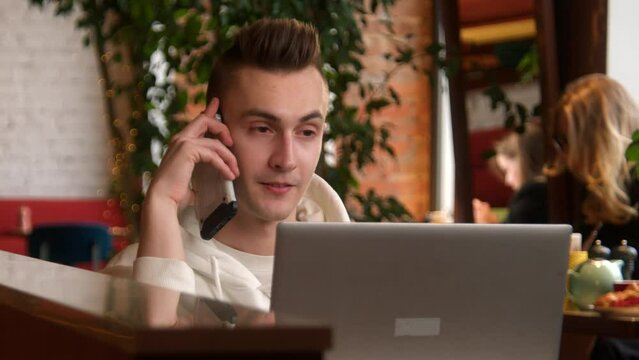 Young man is talking on phone at laptop in cafe. Stock footage. Young man is talking on phone while working on laptop. Man answers work calls while working on laptop in cafe