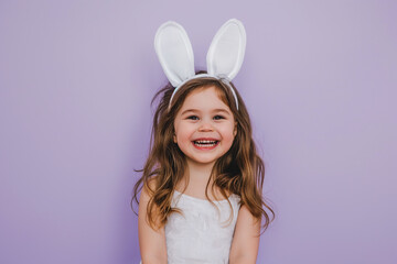 little smiling girl with artificial bunny ears on her head on a purple background, Easter mood