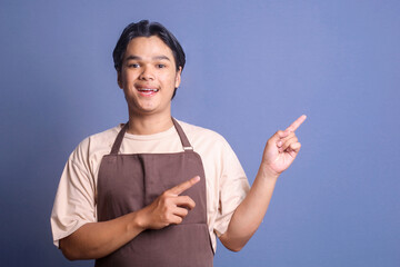 Young Asian man barista employee wearing brown apron working in coffee shop, pointing finger aside...