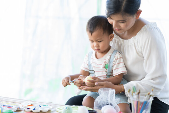 Asian Mother And Baby Little Son Painting Eggs With Brush In Class Workshop. Happy Asian Mom Embracing His Little Son And Teaching Painting Eggs With Water Colour