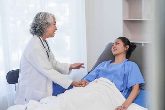 An elderly Asian doctor is smiling and holding the hand of a younger Asian patient who is lying in a hospital bed.