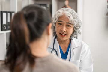 An elderly Asian doctor is talking to a younger Asian woman across a desk in a medical office, monthly health check appointment.