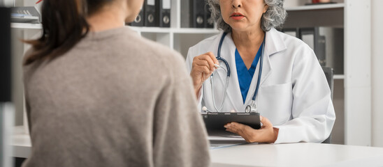 An elderly Asian doctor is talking to a younger Asian woman across a desk in a medical office,...