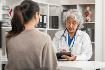 An elderly Asian doctor is talking to a younger Asian woman across a desk in a medical office,...