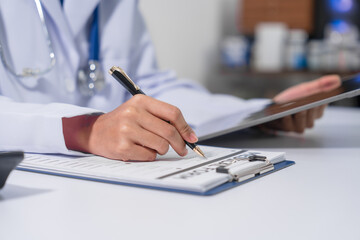 Fototapeta premium A doctor's hand is writing on a medical record form on a clipboard, with a stethoscope and tablet in the background.