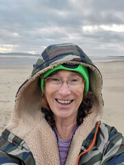 Mature woman takes a selfie after open water swimming. She is wearing layers of warm clothing including a camouflage robe and bobble hat to restore her core temperature.
