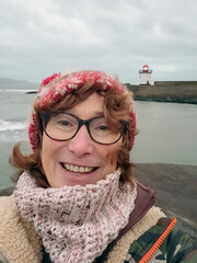 Mature woman takes a selfie at dusk while walking past Burry Port Lighthouse. The weather is overcast and she is wearing a warm camouflage robe and Christmas themed bobble hat.