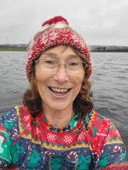 Mature woman wears a colourful rash vest and Christmas themed bobble hat while she swims in a reclaimed dockland on Christmas Day.