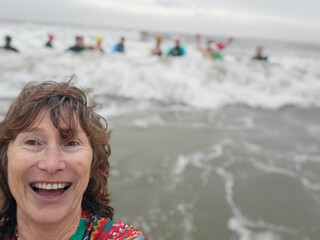 Mature woman wears a colourful rash vest while swimming in the sea on Boxing Day. Other swimmers are out of focus while splashing in the waves.