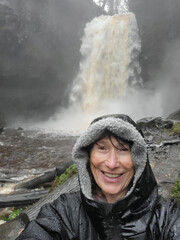Mature woman takes a selfie at Henrhyd Falls after torrential rainfall - one of the many waterfalls in Waterfall Country, near Pontneddfechan in South Wales. She is drenched because of the water spray
