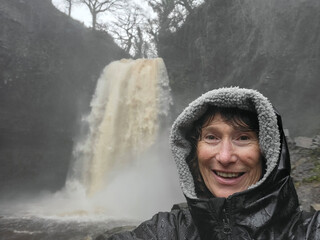 Mature woman takes a selfie at Henrhyd Falls after torrential rainfall - one of the many waterfalls in Waterfall Country, near Pontneddfechan in South Wales. She is drenched because of the water spray