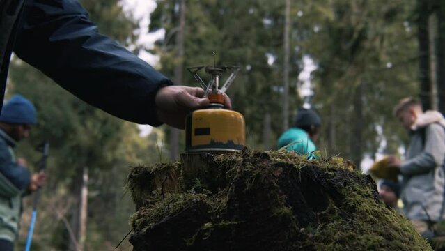 Male hiker sets up portable camping gas stove on stump. Tourist puts kettle on gas burner. Group of hiking buddies stopped to rest during trip or expedition. Active leisure. Slow motion. Close up.