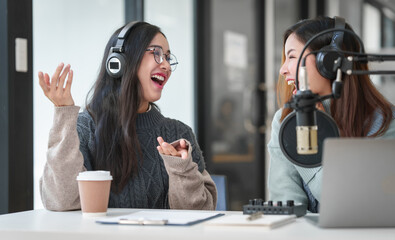 Pretty Asian women podcasting. One speaks into microphone and wears headphones, while the other...