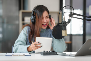 An Asian woman is podcasting alone, speaking into a microphone, wearing headphones, and adjusting...