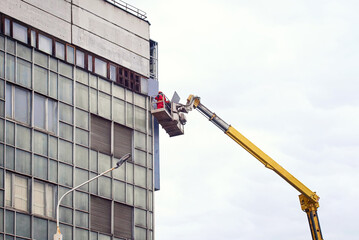Man on aerial platform installing advertising LED panel on facade wall of the building. Worker in crane bucket work at height. Men in crane basket repair outdoor advertising LED display screen
