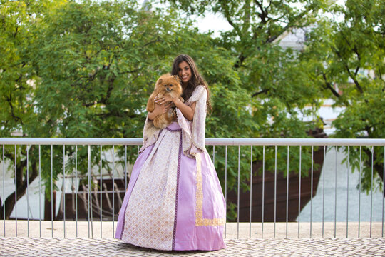 Beautiful Latin woman with long curly hair wearing a 15th century dress is holding a pomenarian dog in her arms. The woman is happy with her pet. Dog and pet day concept.