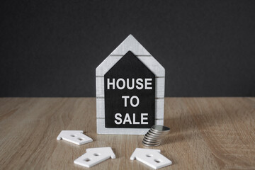 House to sale symbol. Black board in the shape of a house with words house to sale near miniature house. Beautiful wooden table, black background copy space. Business and house to sale concept.