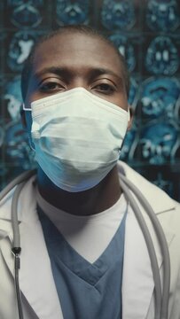 Vertical chest up portrait of young Black male surgeon looking at camera and pulling up face mask while standing against patient brain x-ray in background