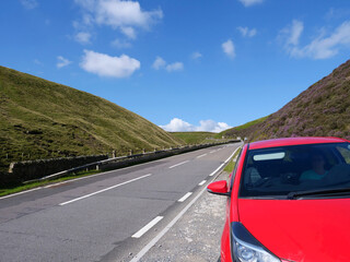Unrecognizable woman sitting inside of red car parked on roadside of winding between hills Snake Road or Snake Pass in Peak District National Park of United Kingdom.
