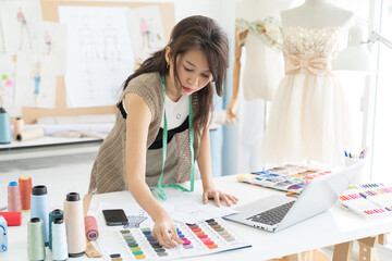 Asian female dressmaker sketching fabric dress and design in sewing workshop. Tailor shop. Garment,...